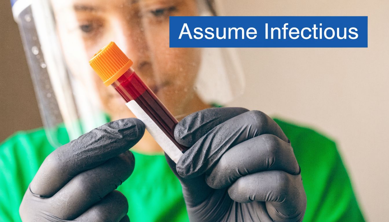 A medical worker wearing a face shield and black gloves holds a blood sample tube labeled assume infectious.