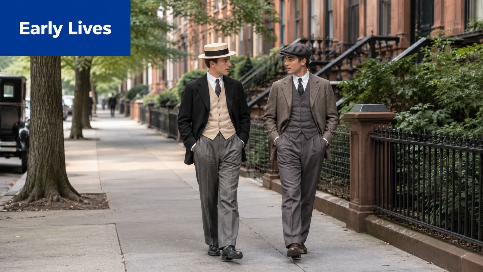 Two men dressed in vintage 20th-century suits walking together along a sidewalk lined with brownstone houses.
